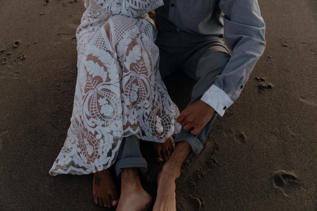 A couple and their intertwined legs sitting on the beach in Oregon at sunset in their wedding attire 